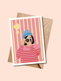 Carte postale Clic de Cœur avec enveloppe kraft, illustration pop d’une photographe en bonnet bleu et fond rose rayé.